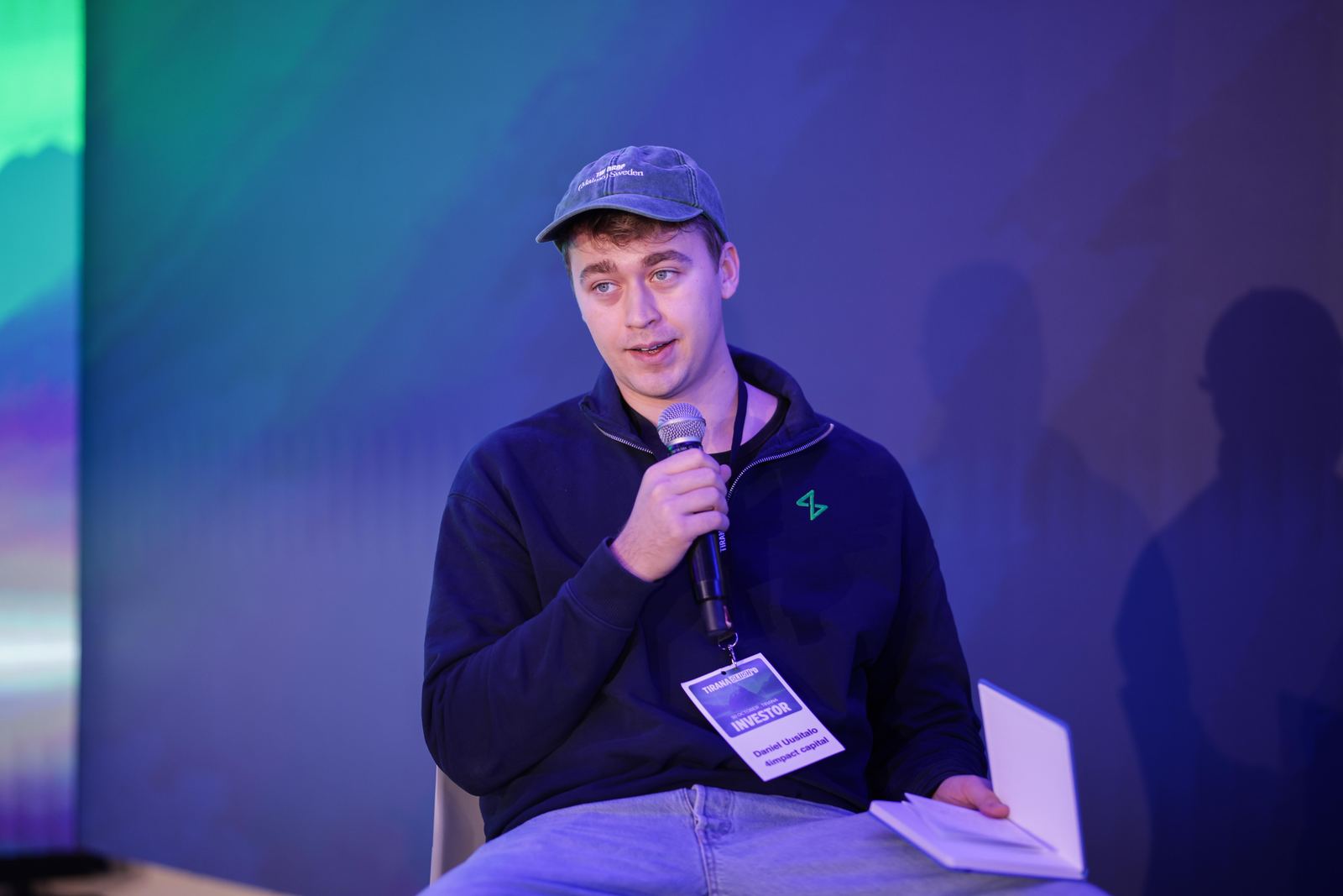 Daniel Uusitalo speaking on stage with a handheld microphone and an investor badge at Tirana Slush'd in Albania